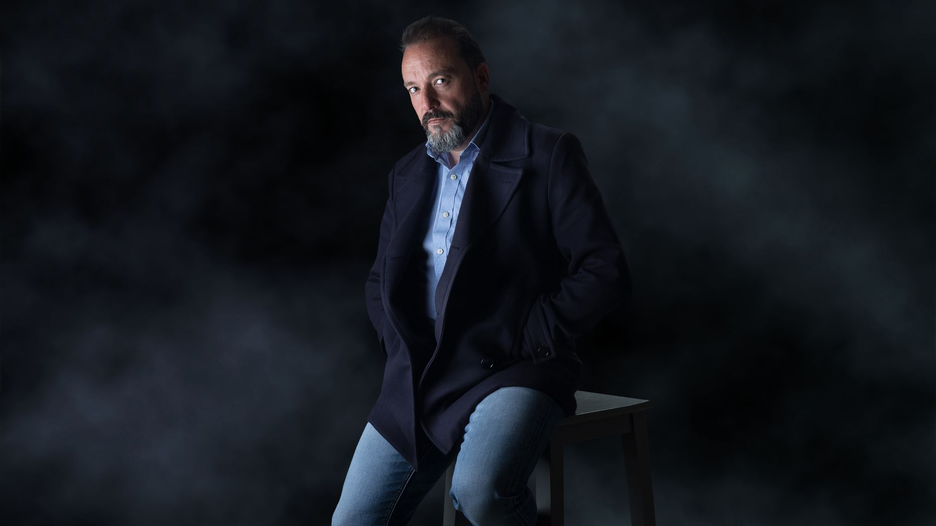 Man wearing dark indigo jeans with a button-down shirt and navy blazer in an office
